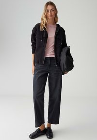 Felpa nera con zip, due tasche frontali, t-shirt rosa chiaro sotto, jeans neri larghi e sneakers nere, tenuta con una borsa nera.