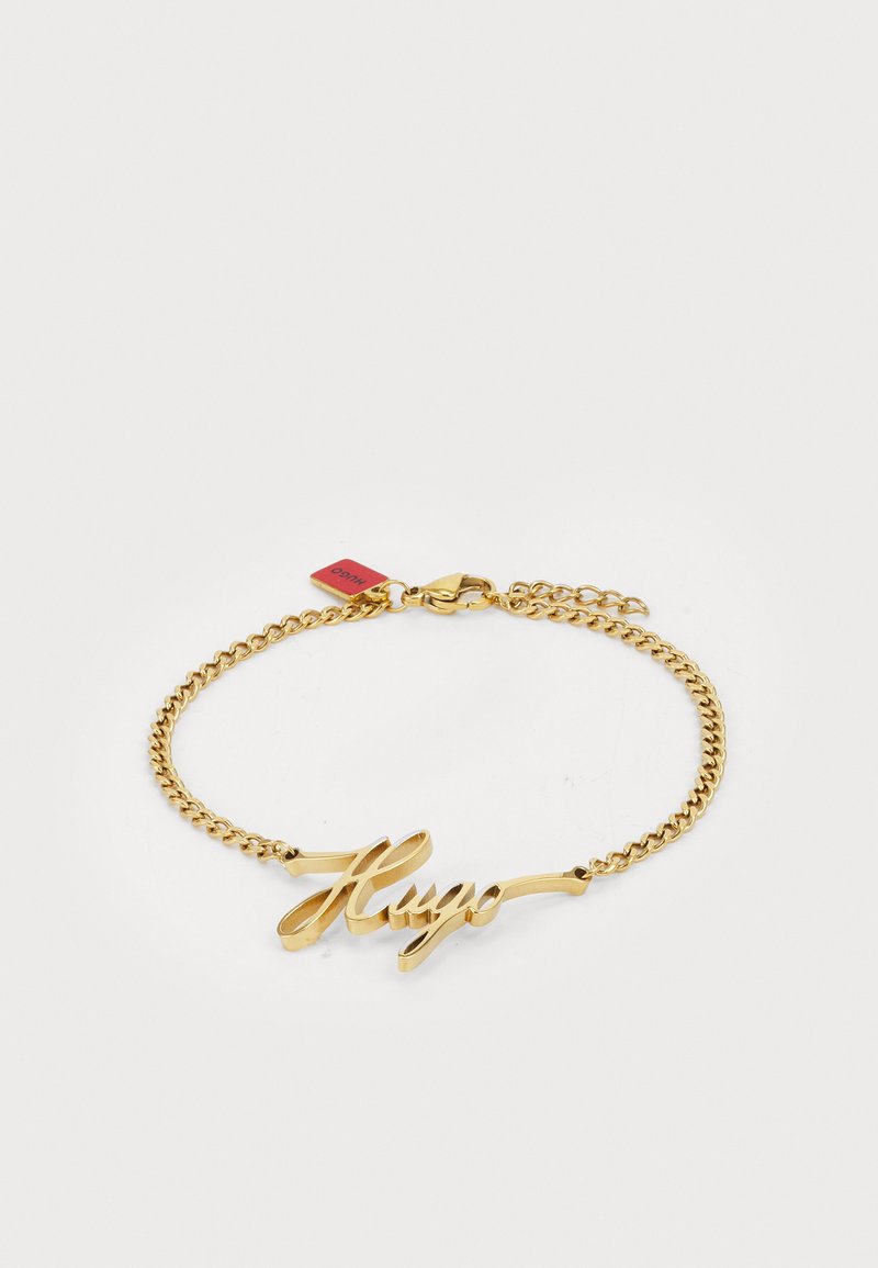 Bracelet en chaîne dorée avec une plaque ornée en cursive gravée "Hugo". Longueur ajustable avec un fermoir et une étiquette.