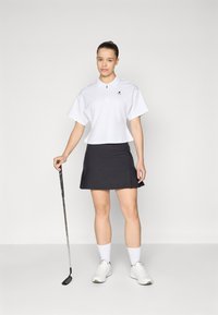 Balta trumpa polo marškinėliai su apykakle, derinami su tamsiu klostuotu golfo sijonu. Modelis laiko golfo lazdą, vilkėdamas baltas kojines ir batelius.