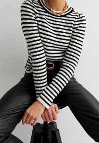 Pull rayé noir et blanc avec texture côtelée, col rond et accents en chaîne dorée sur les épaules, associé à un pantalon noir.