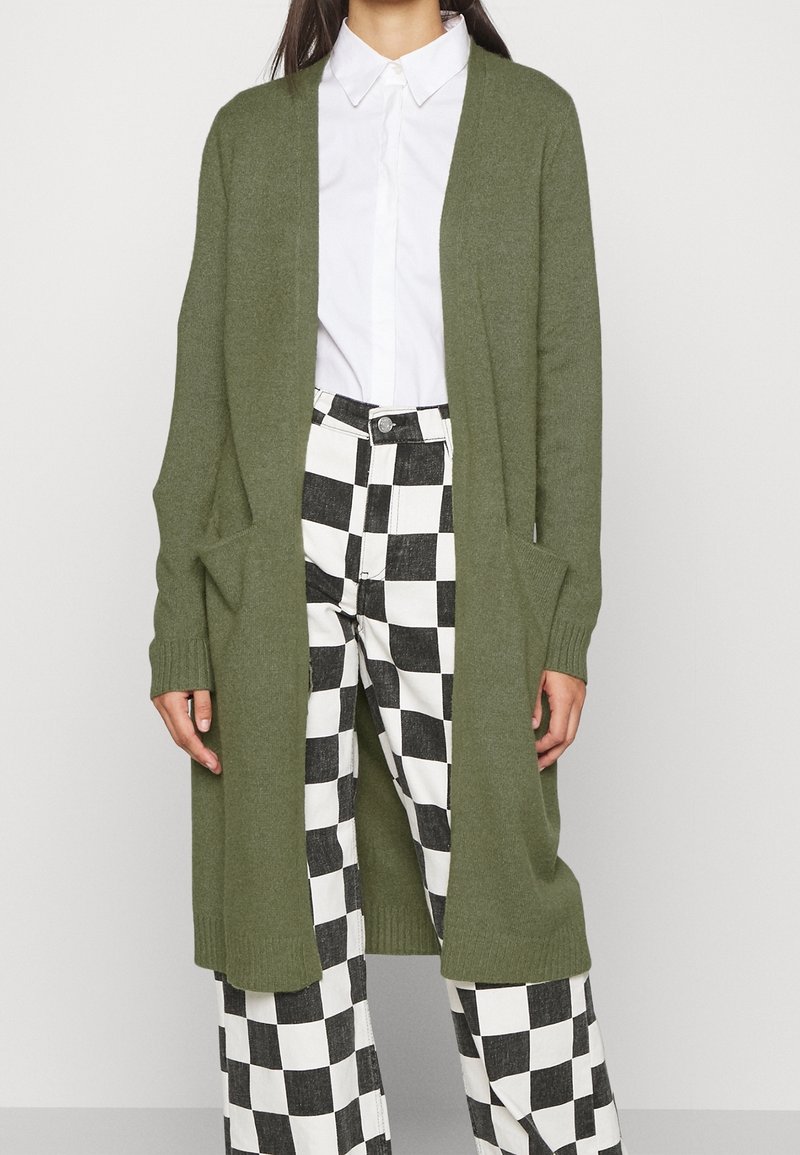 Vrouw in een lange olijfgroene cardigan over een witte button-up shirt en zwart-witte geruite wijde broek.