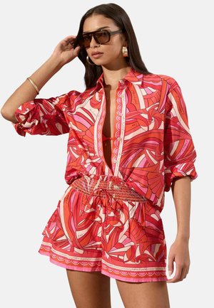 Kobieta nosząca oversize koszulę na guziki w czerwono-różowo-białe wzory, z pasującymi szortami, dużymi okularami przeciwsłonecznymi, kolczykami kołami i złotą bransoletką.