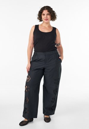 Femme aux cheveux bouclés debout, les mains dans les poches, portant un haut noir sans manches et un pantalon large avec des détails en dentelle, des ballerines noires.