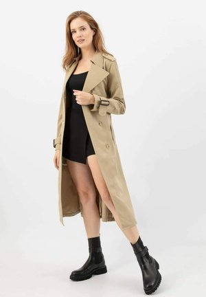 Beige trenčkot s opaskem a velkými knoflíky, spárovaný s černým bodysuitem a černými kotníkovými botami, stojící na bílém pozadí.