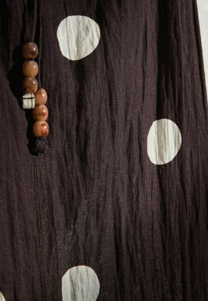 Dunkelbrauner Stoff mit großen weißen Punkten und einer vertikalen Schnur aus fünf braunen Perlen, die nahe am linken Rand angebracht ist.