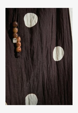 Dunkelbrauner Stoff mit großen weißen Punkten und einer vertikalen Schnur aus fünf braunen Perlen, die nahe am linken Rand angebracht ist.