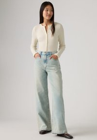 Κρεμ ριγέ μπλούζα με μακριά μανίκια και κουμπιά για κλείσιμο, σε συνδυασμό με ανοιχτό μπλε τζιν παντελόνι φαρδύ και μαύρα επίπεδα παπούτσια, σε ουδέτερο φόντο.