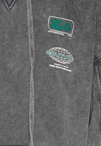 Sudadera gris oscuro con escote en V y tejido texturizado. Presenta tres logotipos en colores contrastantes: detalles en verde y blanco.