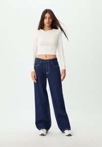 Bjela duga rukava skraćena majica u kombinaciji s tamnoplavim širokim trapericama s laganim šavovima i prednjim džepovima. Model nosi bijele tenisice.