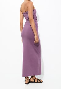 Robe maxi sans bretelles en violet, présentant un détail froncé sur le côté. Assortie à des sandales noires à lanières. Tissu lisse avec une coupe ajustée.