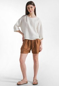 Ung kvinde stående med hånden på hoften iført hvid løs top, brune shorts og beige sandaler mod en ensartet lys baggrund.