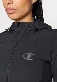 Melna sporta jaka ar rāvējslēdzēju un kapuci, ar spīdīgu logotipa akcentu uz krūtīm un regulējamiem aukliņām. Gluda, viegla auduma.