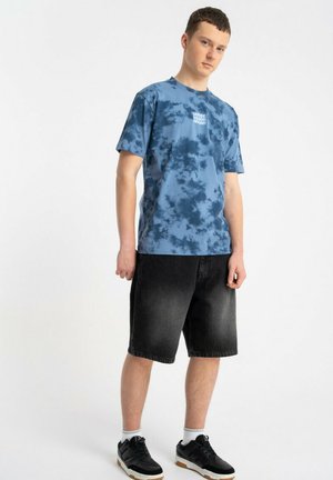 Młody mężczyzna stojący, ubrany w niebieską koszulkę w stylu tie-dye, czarne krótkie spodenki do kolan, białe skarpetki i czarne tenisówki na białym tle.