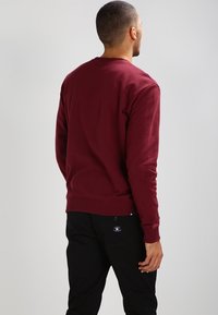 Vinröd sweatshirt med rund halsringning och ribbade ärmslut, bärs tillsammans med svarta byxor. Tyget verkar slätt och av medelvikt.