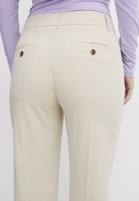 Pantaloni beige su misura con tasche posteriori e bottoni marroni. Tessuto morbido con una texture liscia, dal design a gamba dritta.