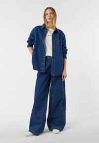 Koszula jeansowa w ciemnoniebieskim kolorze, zapinana na guziki, z kieszenią na piersi; zestawiona z szerokimi spodniami jeansowymi w ciemnym niebieskim odcieniu oraz kremowymi wsuwanymi butami.