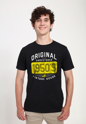 Ung mand med krøllet hår smilende, iført sort t-shirt med gul "1950'erne" og hvid vintage tekst, stående mod en ensfarvet hvid baggrund.
