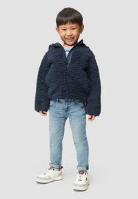 Marineblauwe, fuzzy fleece jas met een rits aan de voorkant, gecombineerd met lichtblauwe jeans en witte sneakers met zwarte en rode accenten.