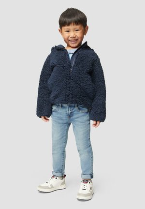 Marineblauwe, fuzzy fleece jas met een rits aan de voorkant, gecombineerd met lichtblauwe jeans en witte sneakers met zwarte en rode accenten.