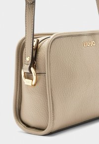 Beige leren schoudertas met een ronde vorm, gouden hardware, ritsafsluiting en een merkembleem, voorzien van textuurdetails.
