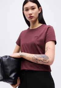 Νεαρή γυναίκα με μακριά σκούρα μαλλιά φοράει μπορντό t-shirt BOSS, μαύρο παντελόνι, κρατά μια μαύρη δερμάτινη τσάντα και δείχνει ένα λεπτομερές τατουάζ στο μπράτσο της.