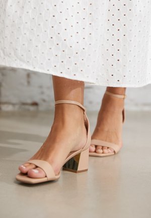 Voeten met nude blokhak sandalen met enkelbandjes, gecombineerd met een witte midi rok met opengewerkt motief, staand op een gladde lichte vloer.
