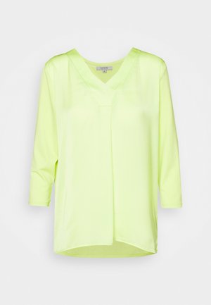 Blouse jaune clair à manches longues avec col en V et détail de pli devant, présentée sur un fond blanc uni.