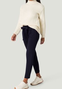 Femme portant un pull en tricot crème, un pantalon taille haute en papier bleu marine avec un nœud, et des baskets blanches, se tenant devant un fond clair.