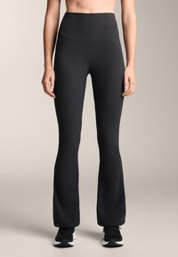 Fekete, bővülő szárú, aktív sportruházati leggings magas derékpánttal, sima, rugalmas anyagból készült, fekete sportcipővel párosítva.