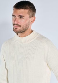 Cremfarbener, gerippter Strickpullover mit hohem Kragen und legerer Passform. Verfügt über einen strukturierten Stoff und dezente Nähte entlang der Schultern.