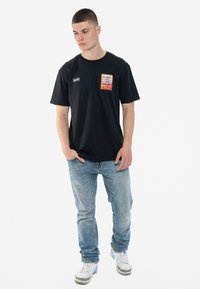 Black short-sleeved t-shirt with a chest pocket featuring a colorful graphic label. Worn with light blue jeans and sneakers.