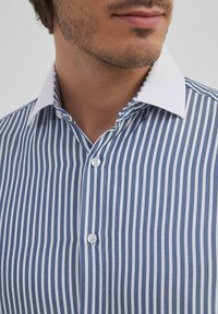 Chemise à rayures blanches et bleues avec un col blanc bien net, présentant une fermeture à boutons et un tissu texturé. Coupe décontractée, sans motifs.