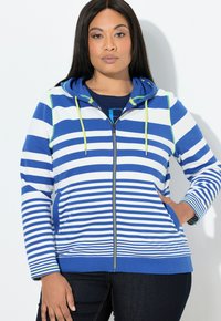 Žena v modro-bílé pruhované bundě na zip s limetkově zelenými šňůrkami a tmavých džínách, stojící před bílým pozadím.