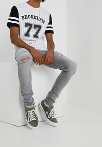 Hombre vistiendo una camiseta blanca y negra con "Brooklyn 77", jeans grises rasgados y zapatillas verdes, sentado en un bloque blanco contra una pared blanca.