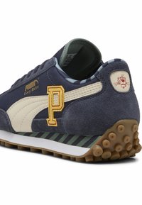 Zapatillas de ante azul con acentos beige, que presentan un logo "P" amarillo, suela rayada y suela exterior de goma texturizada. Diseño retro con branding.