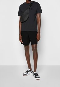 Man som bär en svart OBEY t-shirt, svarta shorts, en svart crossbody-väska och svart-vita sneakers, stående mot en enfärgad vit bakgrund.