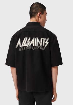 Man trägt ein schwarzes kurzärmeliges Shirt mit großem weißem Text „ALLSAINTS SEE THE UNSEEN“ auf dem Rücken und steht vor einem einfarbigen hellen Hintergrund.