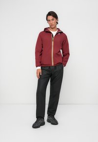 Chaqueta ligera color marrón con cremallera y capucha amarillas, combinada con pantalones negros. Zapatillas oscuras completan el look contra un fondo neutro.