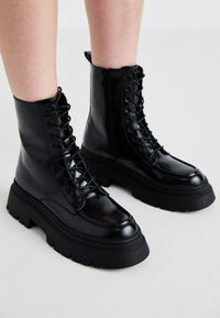 Bottines en cuir noir avec une semelle épaisse, un devant lacé et une fermeture éclair sur le côté ; présente une finition lisse et un embout surélevé.