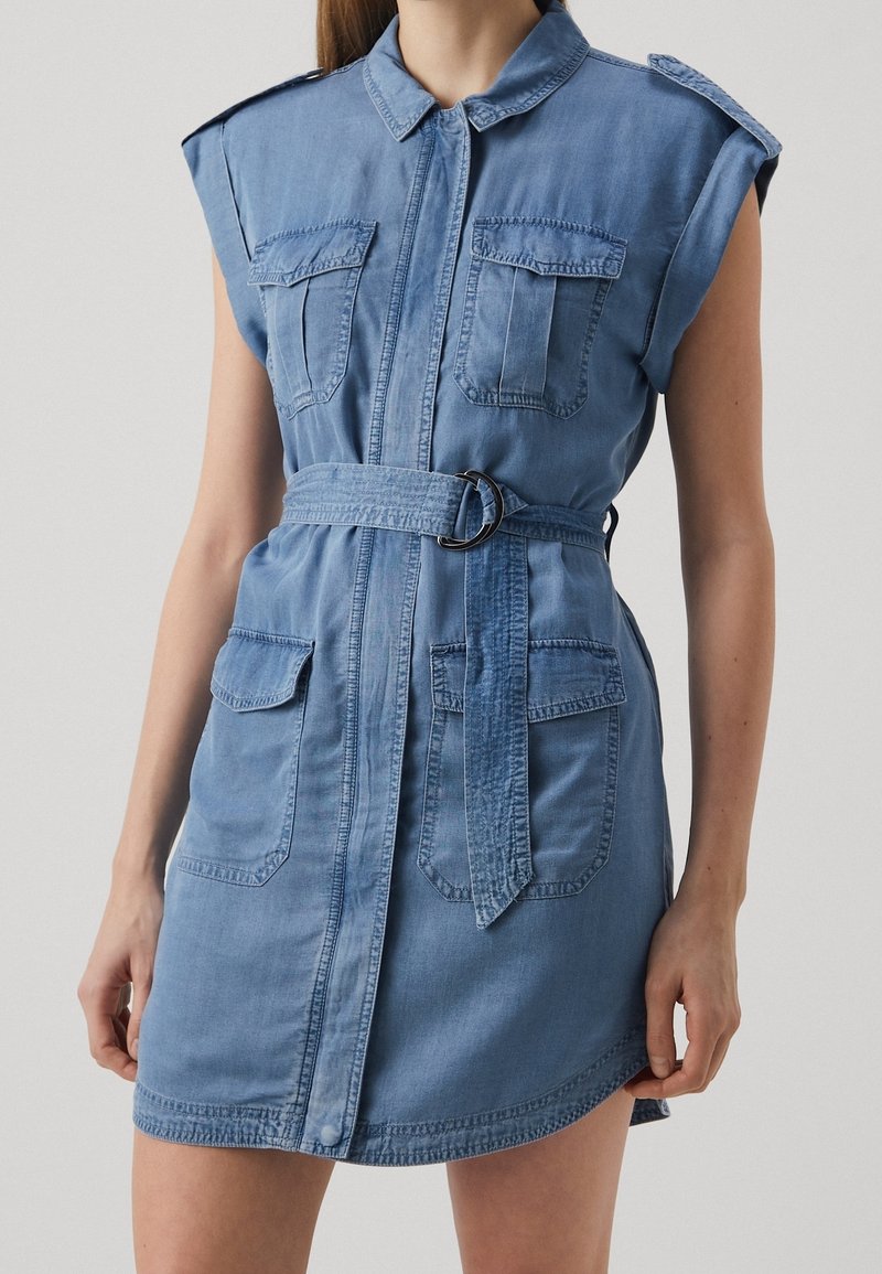 Robe chemise en denim avec manches courtes, deux poches poitrine, une taille ceinturée et un col, de couleur bleue claire avec une texture subtile.