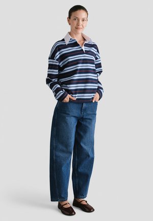 Kobieta stoi z rękami w kieszeniach, ubrana w niebieską w miejscu w paski, długą koszulę, szerokie dżinsy oraz ciemnobrązowe płaskie buty na tle jednolitego tła.