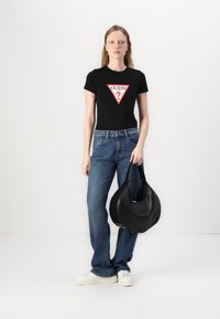 T-shirt noir ajusté avec le logo triangulaire rouge "GUESS?", associé à un jean bleu et un sac à épaule en cuir noir. Des baskets blanches complètent le look.