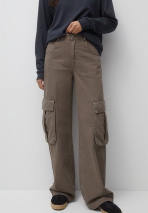 Personne portant un pantalon cargo marron à jambes larges avec de grandes poches latérales et des chaussures noires sans lacets, debout devant un fond clair uni.