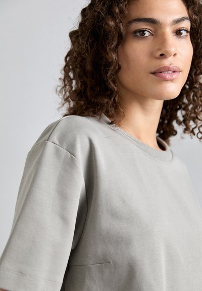 Ανοιχτό γκρίζο κοντομάνικο μπλουζάκι από μαλακό βαμβάκι, με στρογγυλή λαιμόκοψη και λεπτομέρεια τσέπης. Λείο ύφασμα.