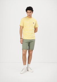 Tricou cu mânecă scurtă, în dungi galbene și albe, cu un logo mic, asortat cu șorturi verzi și adidași albi. Design simplu, croi lejer.