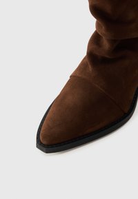 Bottine en suede marron avec un bout pointu et un design décontracté. Dotée d'une semelle en caoutchouc noire et de coutures visibles le long du bord.