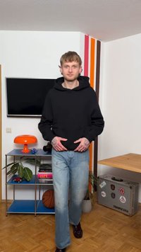 Νεαρός άνδρας με μαύρο φούτερ με κουκούλα και τζιν στέκεται σε δωμάτιο με ρετρό πορτοκαλί λάμπα, μπάλα του μπάσκετ, βιβλιοθήκη, γλάστρες και βαλίτσα με την ετικέτα "TROPICANA".