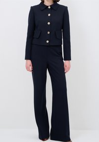 Mörkblå kostym med en kraghängd jacka som har detaljer i guldknappar och två framfickor, parat med vida byxor. Slät tygtextur.