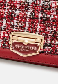 Červená a černá tkanina se zlatým zapínáním. Obsahuje zlatě rytou destičku se nápisem "RIVER ISLAND LONDON" připevněnou k červenému podkladu.