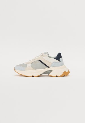 Casual sneaker met beige, grijze en marineblauwe panelen, witte veters en een dikke witte zool met bruin profiel op een effen achtergrond.
